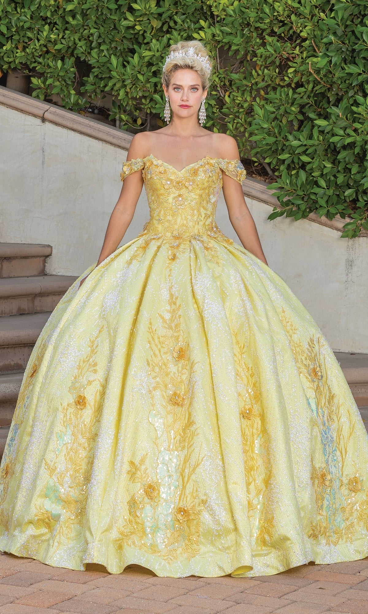 A woman stands outdoors on stone steps in front of greenery and a stone wall, wearing the Quinceanera Dress 1772 By Dancing Queen—an off-shoulder A-line ball gown with gold embroidery and a sweetheart neckline, paired with a tiara.